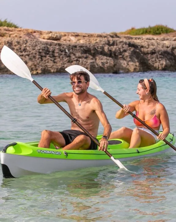 Due persone sorridenti su un kayak verde che remano insieme in acque limpide vicino a una costa rocciosa.