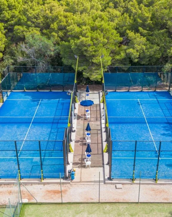 Due campi da paddle blu affiancati con cabine ombrelloni e sedie bianche, circondati da alberi verdi.
