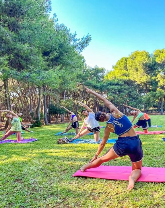 Gruppo di persone che pratica yoga all'aperto su tappetini colorati in un parco con alberi e cielo sereno.