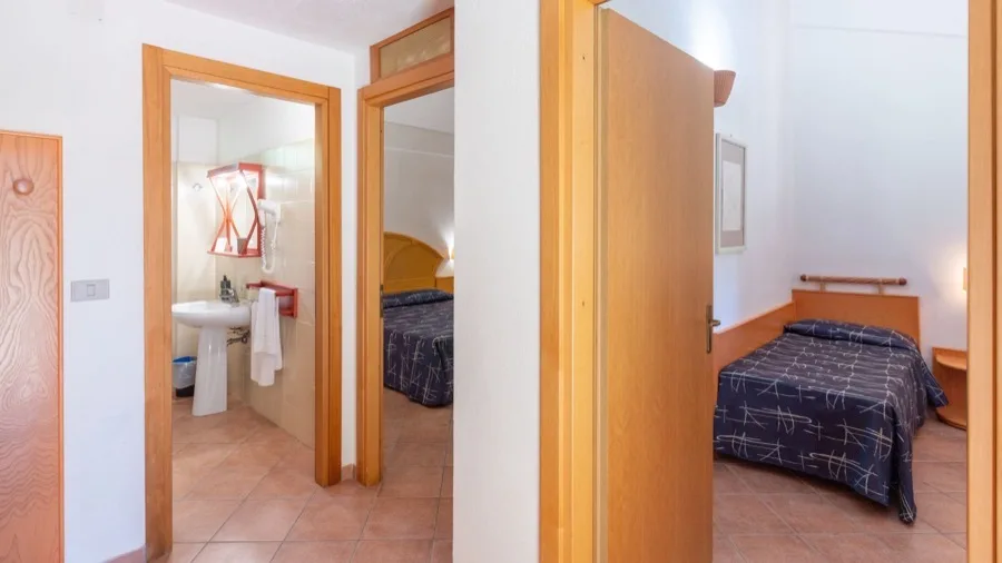 Camera da letto con letto singolo e bagno con lavabo e specchio, porta in legno aperta su pavimento in piastrelle.