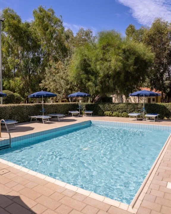 Piscina all'aperto con lettini e ombrelloni blu, circondata da siepi e alberi verdi sotto cielo azzurro.
