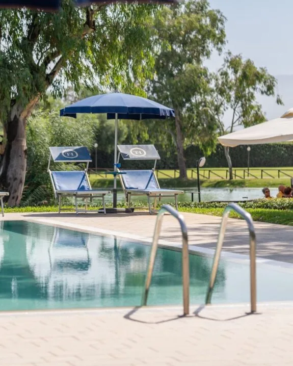 Piscina all’aperto con due sdraio blu sotto ombrellone e alberi verdi sullo sfondo.