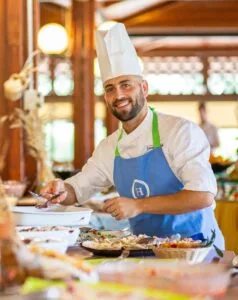 Cuoco sorridente con cappello e grembiule blu prepara piatti su un bancone in ambiente rustico e luminoso.