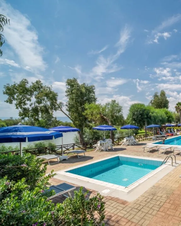 Piscina all'aperto con ombrelloni blu, lettini e sedie bianche, circondata da alberi e cielo azzurro.
