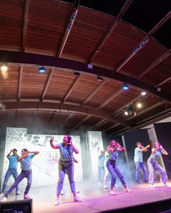 Gruppo di giovani che ballano in abbigliamento casual su un palco illuminato con schermo e fumo scenico.
