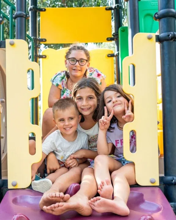 Quattro bambini sorridenti su uno scivolo colorato in un parco giochi all'aperto.