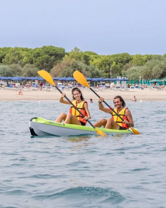 Due persone sorridenti con giubbotti di salvataggio arancioni remano in kayak verde nel mare vicino a una spiaggia affollata.