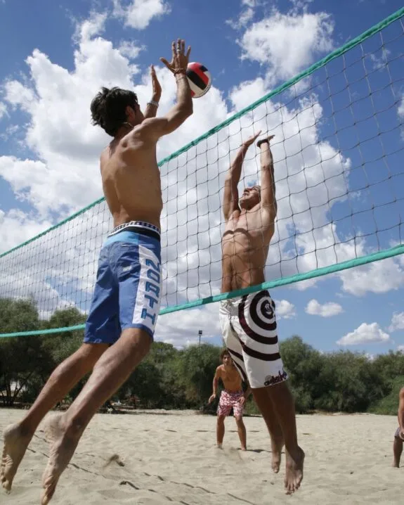 Due uomini senza maglietta giocano a beach volley, uno salta per colpire la palla sopra la rete su sabbia e cielo sereno.