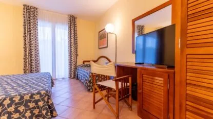 Camera d'hotel luminosa con due letti singoli, scrivania in legno con TV, sedia pieghevole e finestra con tende floreali.