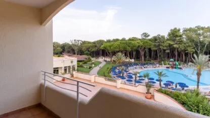 Vista dalla terrazza di un hotel su piscina con ombrelloni blu, alberi e sentiero circondato da vegetazione.
