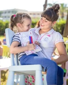 Bambina seduta su sedia, che mostra un disegno a una donna sorridente in ambiente all'aperto.