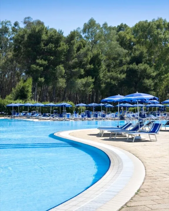 Piscina con lettini e ombrelloni blu, circondata da alberi verdi sotto cielo azzurro.