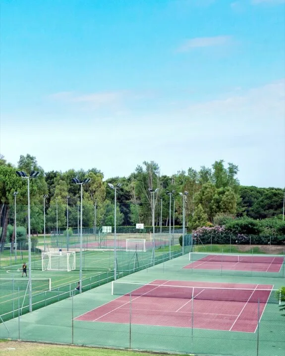 Campi sportivi all'aperto con campi da tennis rossi e campo da calcio verde, circondati da alberi e recinti.