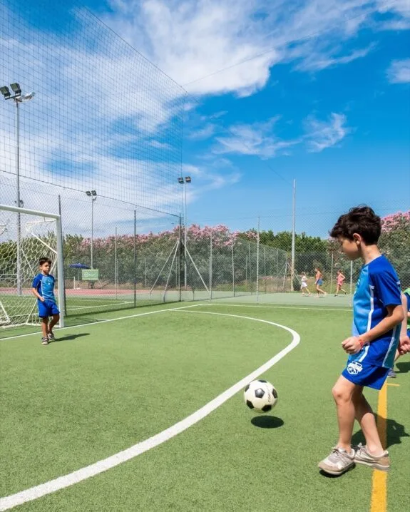 Ragazzo in divisa blu che calcia un pallone da calcio su campo all'aperto con porta e altro bambino in porta.