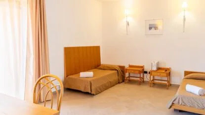 Camera con due letti singoli, comodini in legno, sedia in vimini e tende beige, illuminata da luci da parete.
