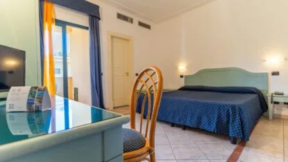 Camera d'hotel luminosa con letto matrimoniale, sedia in legno, scrivania con tv e porta finestra con tende blu e gialle.