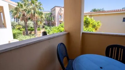 Balcone con tavolo e sedie di plastica blu, vista su palme, edificio rosa e cielo sereno.
