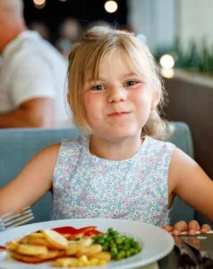 Bambina sorridente con vestito azzurro a fiori mentre mangia patatine, piselli e mais in un ristorante.