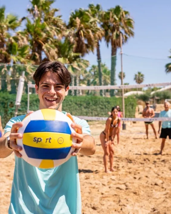 Ragazzo sorridente che tiene un pallone da beach volley su un campo sabbioso con persone e palme sullo sfondo.