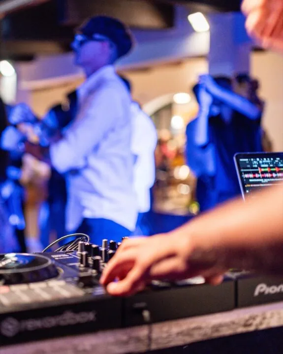 DJ con console mixer e laptop, persone che ballano sfocate sullo sfondo in un ambiente con luci blu.