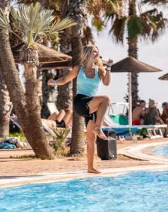 Donna con microfono danza vicino a piscina all'aperto con palme e persone sedute su sdraio al sole.