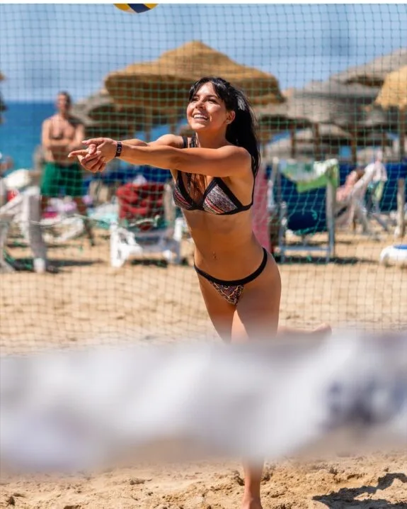 Donna sorridente in bikini colorato gioca a beach volley sulla sabbia con ombrelloni e spettatori sullo sfondo.
