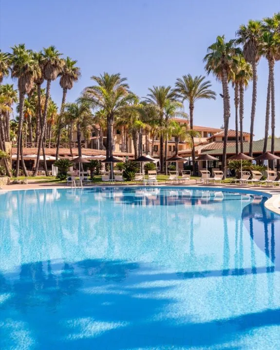 Piscina all'aperto con sdraio e ombrelloni, circondata da palme e edificio in stile mediterraneo.