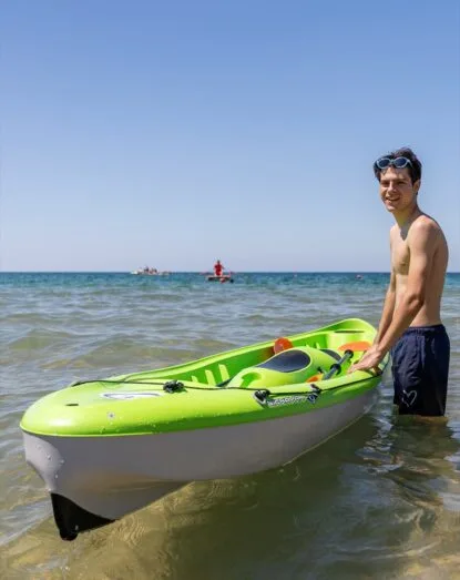 Ragazzo sorridente in piedi in mare accanto a un kayak verde con occhiali da sole sulla testa.