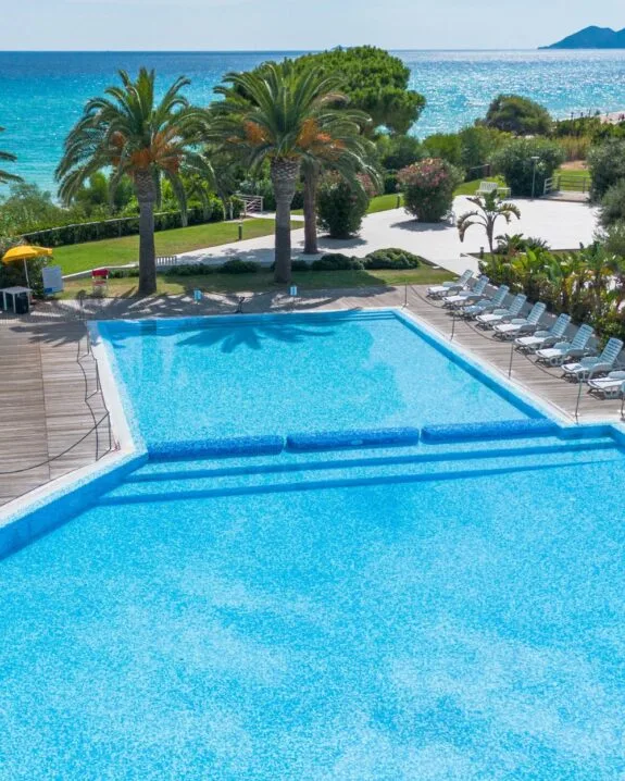Piscina blu circondata da palme, con vista sul mare e lettini a disposizione in un ambiente soleggiato.