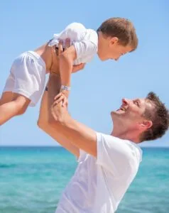 Padre solleva felice il figlio sorridente sulla spiaggia con mare e cielo azzurro sullo sfondo.