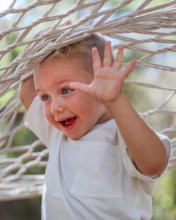 Bambino sorridente con maglietta bianca che gioca tra le corde di un'amaca all'aperto.