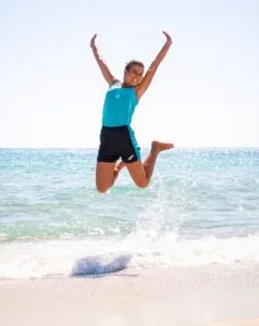 Donna che salta felice in riva al mare con canotta azzurra e pantaloncini neri, con onde e cielo sereno sullo sfondo.