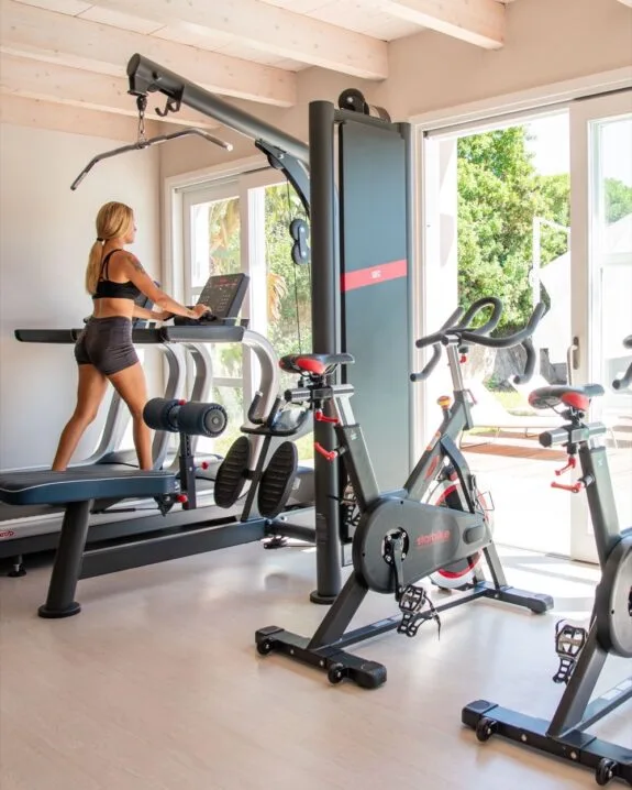 Donna che corre su tapis roulant in palestra luminosa con due cyclette e attrezzi fitness.