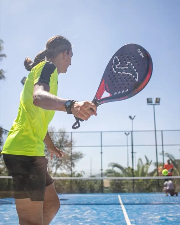 Giocatore di padel in maglia gialla prepara il colpo su campo blu all'aperto sotto cielo sereno.
