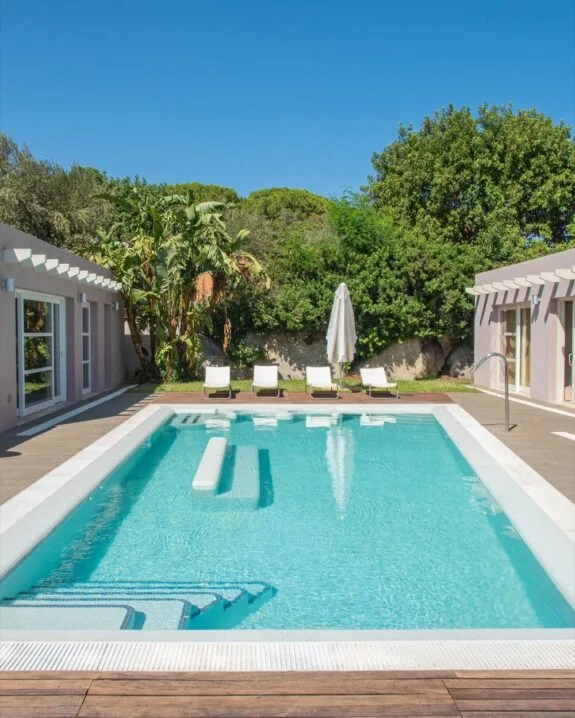Piscina rettangolare con acqua limpida, quattro sdraio bianche e ombrellone, circondata da vegetazione e due edifici.