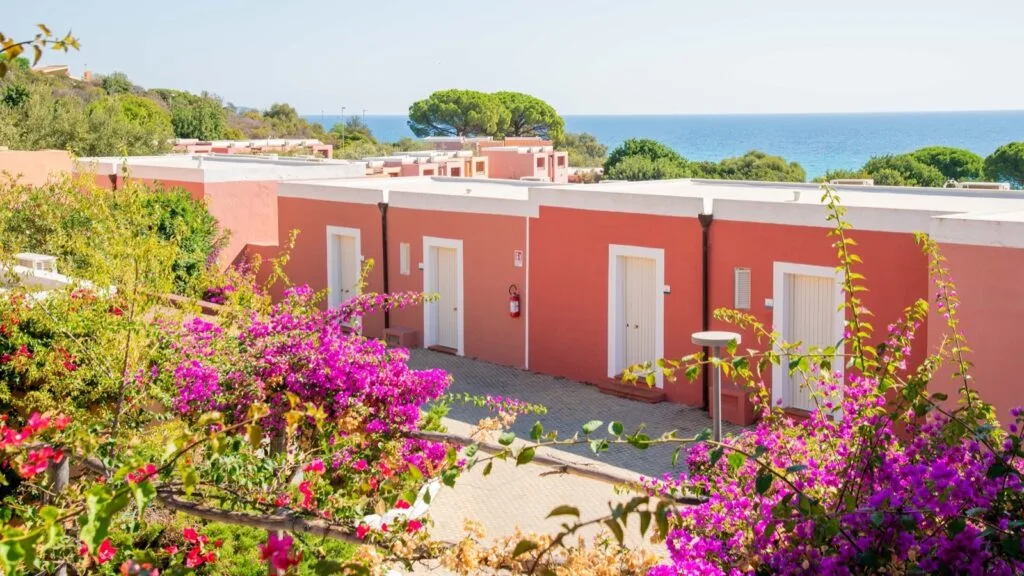 Case rosa con porte bianche, fiori viola in primo piano e vista mare sullo sfondo sotto cielo limpido.