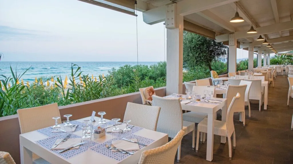 Terrazza di ristorante con tavoli apparecchiati, vista sul mare e ombrelloni gialli sulla spiaggia.
