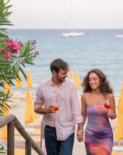 Coppia che cammina mano nella mano sulla spiaggia con cocktail in mano, mare e ombrelloni gialli sullo sfondo.