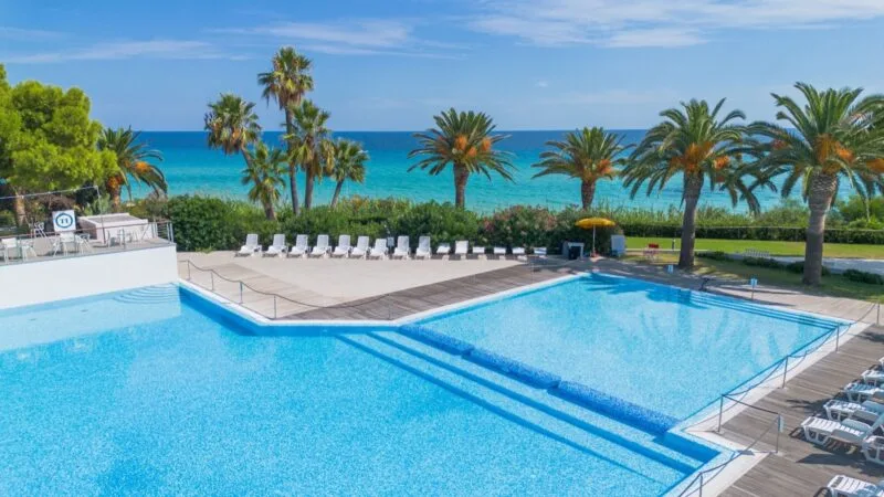Piscina all'aperto con lettini e palme, vista mare e cielo azzurro sereno.