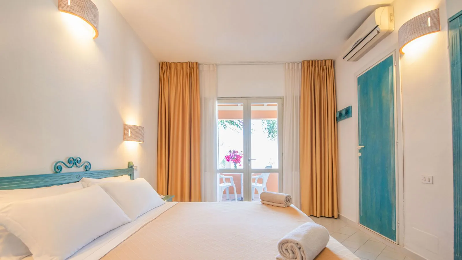 Camera da letto luminosa con letto matrimoniale, tende arancioni, porta finestra e balcone con sedie bianche e fiori rosa.