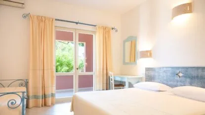Camera luminosa con letto matrimoniale, scrivania azzurra, tende beige e porta finestra che dà su un giardino verde.
