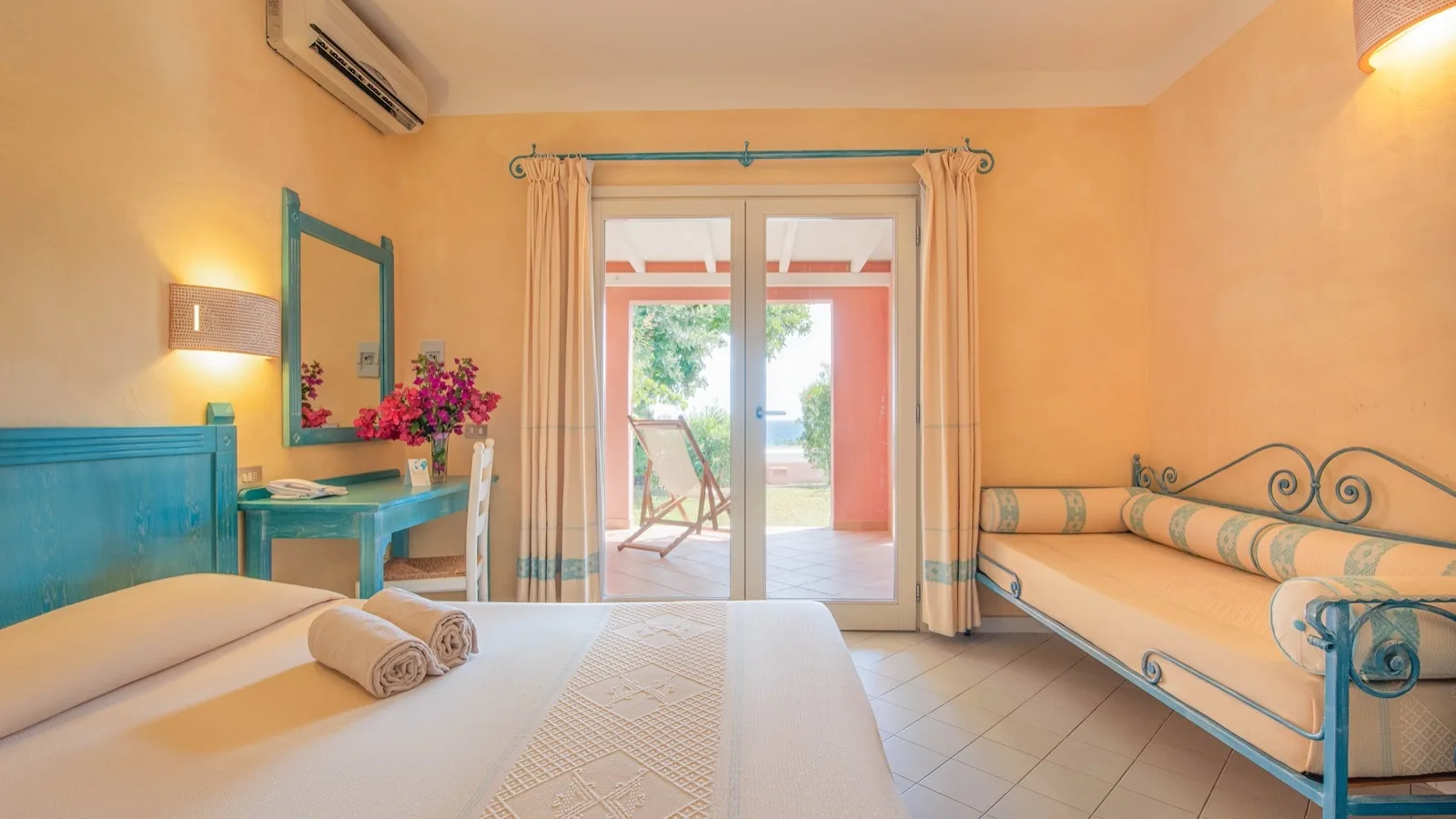 Camera d'hotel luminosa con letto matrimoniale, divano letto, scrivania azzurra e porta finestra su terrazza con vista giardino.