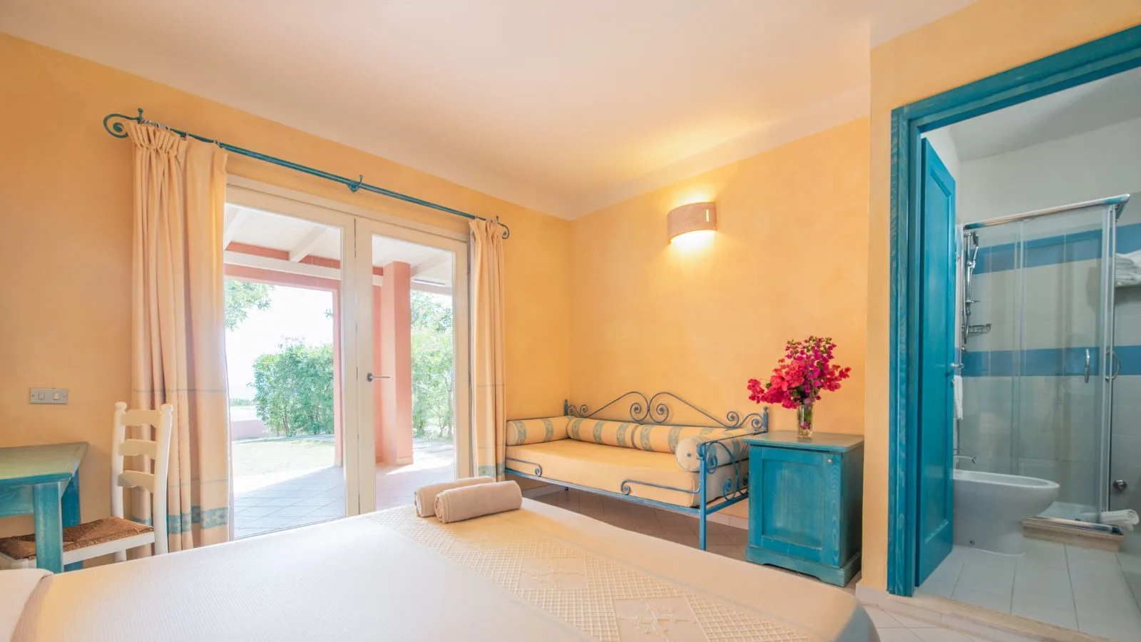 Camera luminosa con letto, divano in ferro battuto blu, armadietto e bagno con doccia, pareti color pesca e balcone.