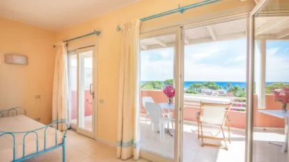 Camera luminosa con letto azzurro, balcone arredato e vista sul mare e vegetazione mediterranea sotto cielo azzurro.