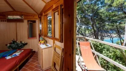 Interno in legno con finestra, vista su alberi e mare, sedia e ombrellone con scritta Touring Club Italiano.