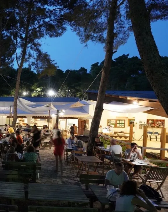 Esterno di un ristorante all'aperto di sera con persone sedute ai tavoli e luci calde, circondato da alberi e natura.