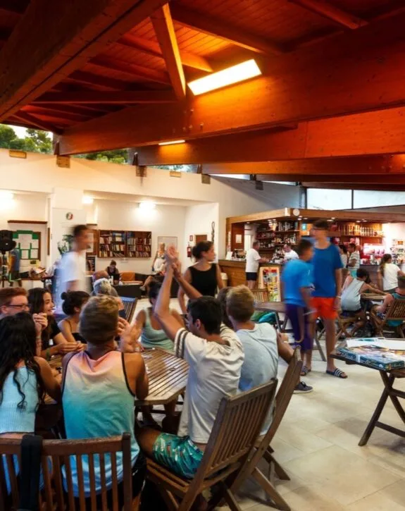 Gruppo di giovani che chiacchierano e giocano a tavola in un ambiente accogliente, con scaffali pieni di libri sullo sfondo.