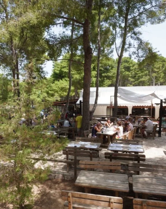 Ristorante all'aperto tra gli alberi con tavoli e persone che mangiano e socializzano sotto un tendone bianco.