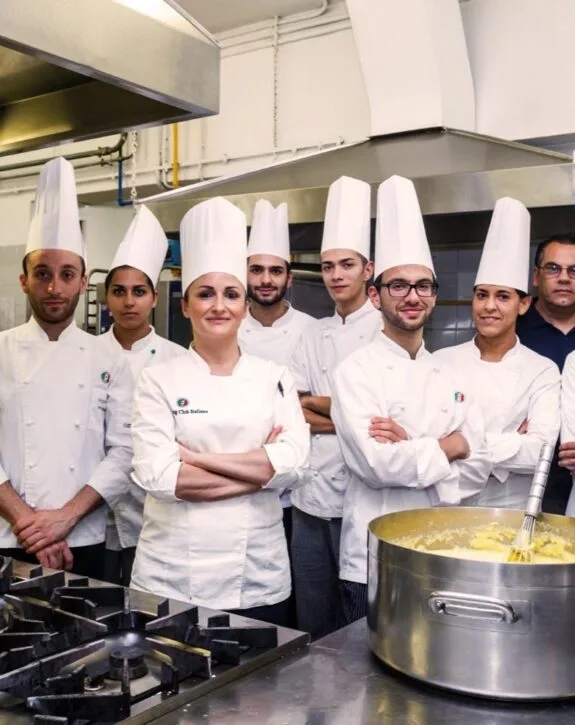 Gruppo di chef in cucina, indossano giacche e cappelli bianchi, pronti a cucinare.