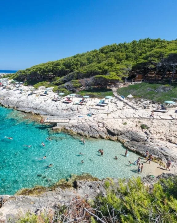 Spiaggia con acque cristalline, persone che nuotano e si rilassano su sedie sotto gli ombrelloni.
